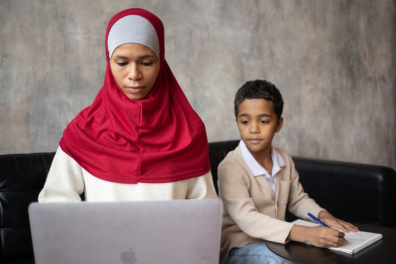 mother in hijab using laptop while son writing