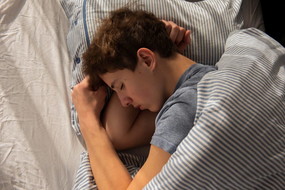 Young man sleeping in bed, lying on his side with a striped blanket around him.