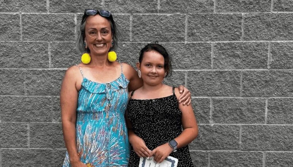 Two smiling women pose arm-in-arm against a gray brick wall; one in a turquoise dress with yellow pom-pom earrings, the other in a black polka-dot dress.